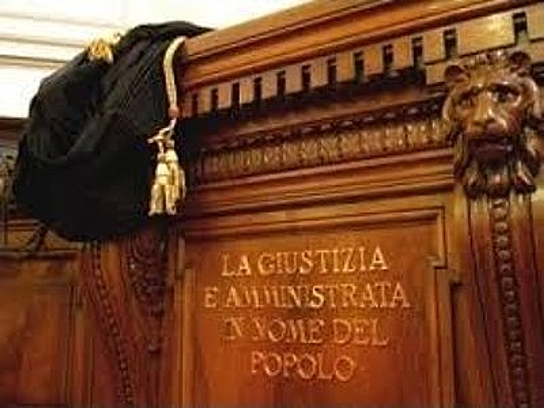 Toga appesa al banco di un'aula di tribunale, sotto l'iscrizione «La giustizia è amministrata in nome del popolo»